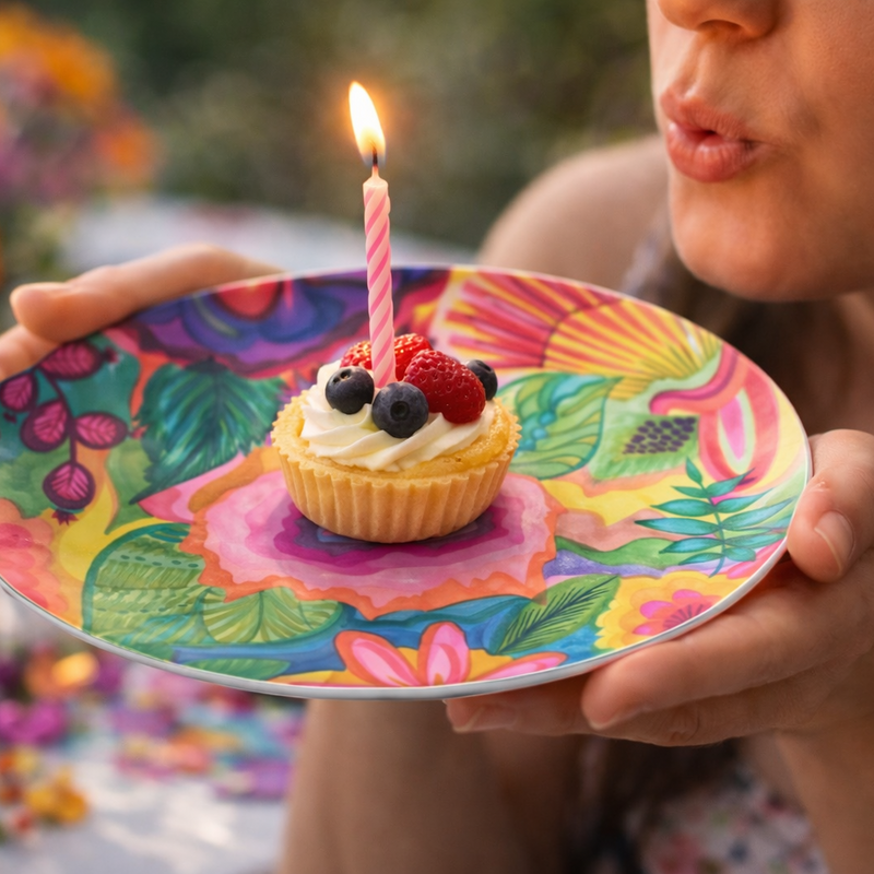 Person blowing out a candle on a small cupcake with berries on a beautiful designer AniVani colorful plate.