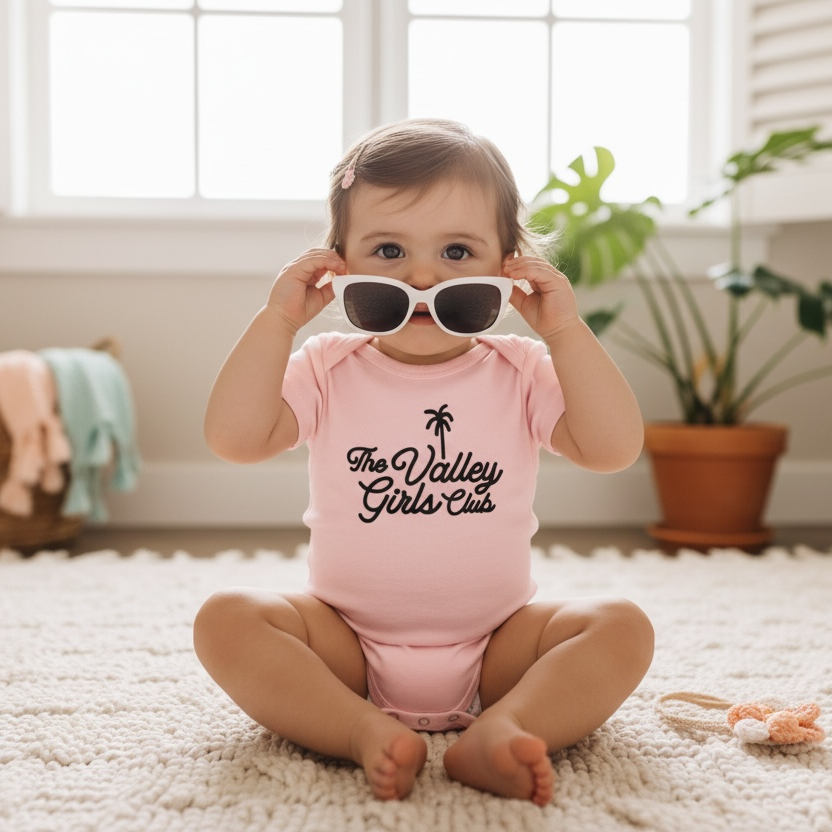 Baby wearing sunglasses and a pink Valley Girls Club onesie with text, sitting on a carpeted floor.