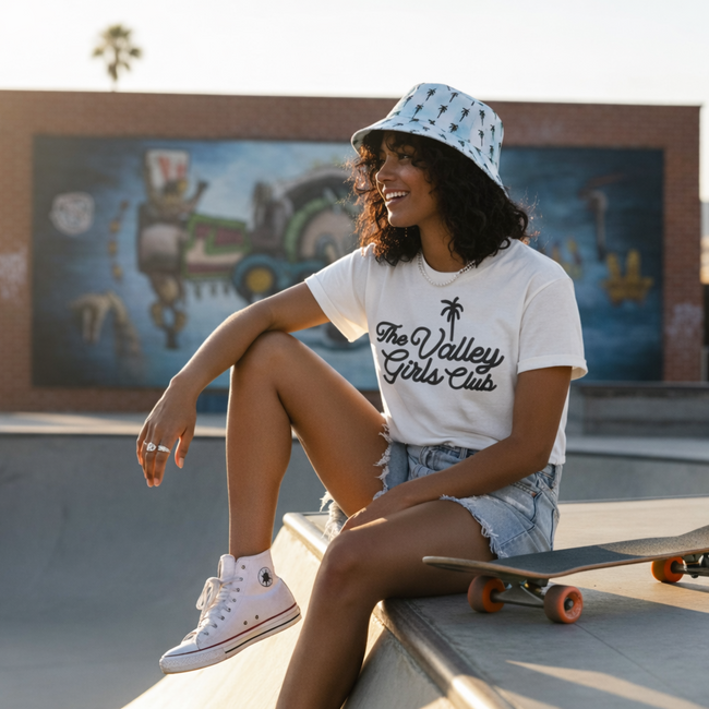 Woman sitting on a skateboard at a skate park wearing a 'The Valley Girls Club' t-shirt and bucket hat