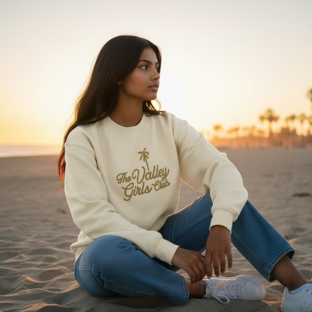 Woman sitting on a beach wearing a cream The Valley Girls Club sweatshirt with text and blue jeans.