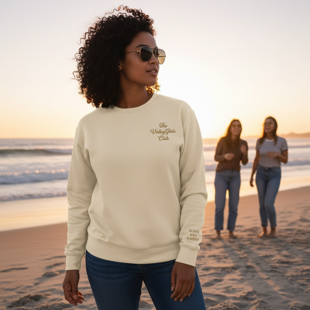 Woman wearing a Valley Girls Club beige sweatshirt with text on a beach at sunset